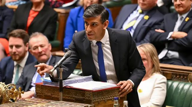 Rishi Sunak, a man, wearing a dark suit and blue tie, speaking into a black microphone in the House of Commons, with his right arm outstretched pointing