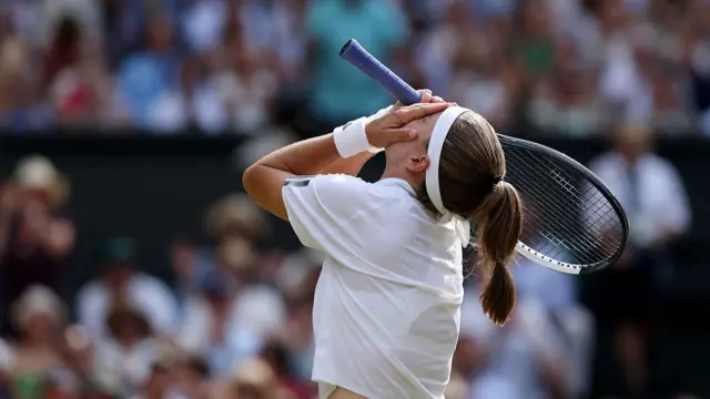 Karolina Muchova, facing away from the camera and wearing a white t-shirt and headband, throws back her head with her hands to her face