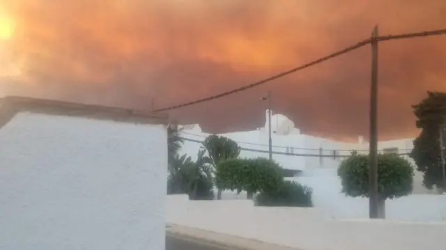 A view of the sky which is burning orange with smoke and clouds in the air hanging above white buildings