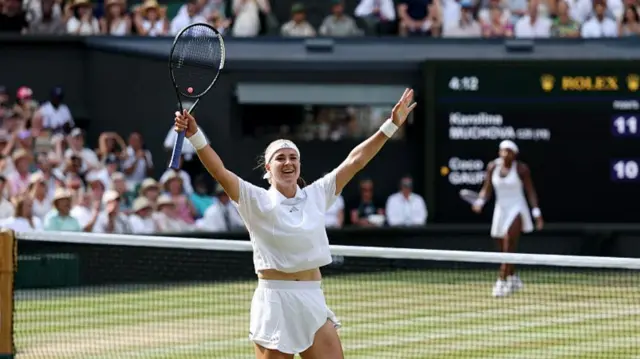 Muchova, wearing a white t-shirt, shorts, and headband, holds her arms outstretched with her racquet in her right hands