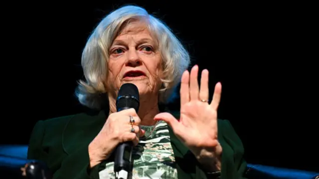 Ann Widdecombe wearing a green blazer and speaking into a black microphone with her left hand outstretched.
