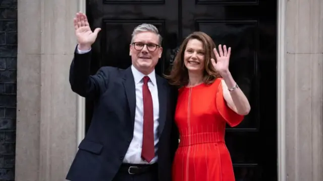 Keir and Victoria Starmer are shown smiling and waving outside the entrance to number 10 Downing Street.