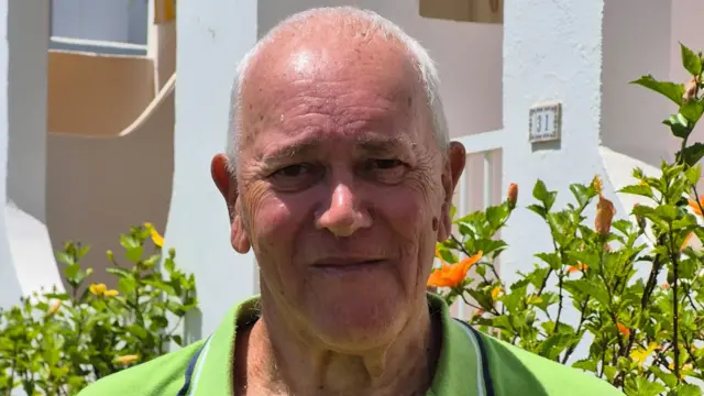 Headshot of Peter who looks at the camera wearing a green shirt, in front of a green bush and white house