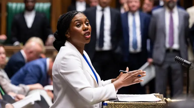 Kemi Badenoch, a woman wearing a white blazer, standing in the House of Commons, looking to her right with her arms in front of her as she talks.