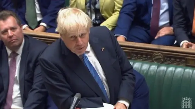 Boris Johnson, a man, wearing a dark suit and blue tie, standing and speaking in the House of Commons with green benches behind him.