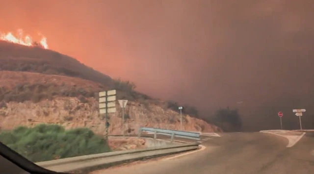 Still from video shows view out of car windscreen as fire rages on hill to left and smoke fills air