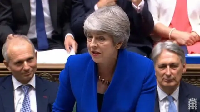 Theresa May, wearing a blue blazer, standing in the House of Commons with two men sitting behind her.