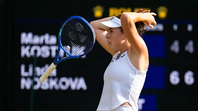 Linda Noskova, wearing a white top and visor, lets go of her racquet and throws her hands behind her head