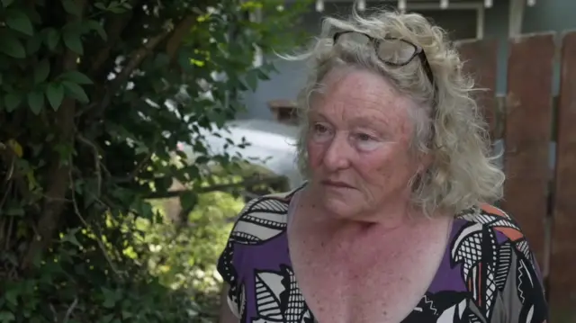 Susan Coysh, a woman, looking to her right as she speaks. Behind her is a green hedge.