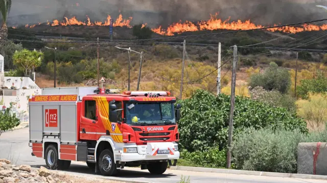 A fire engine in front of a grassy mountain, the fire rages in the distance.