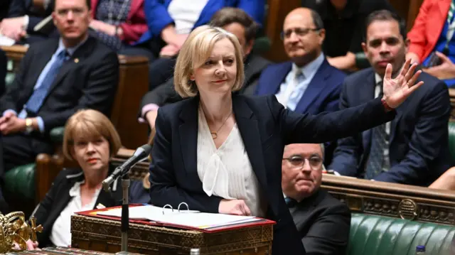 Liz Truss, a woman, wearing a dark blazer, in the House of Commons looking to her left with her arm outstretched