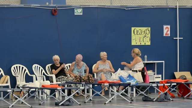 Four women sit in a row on white plastic chairs in front of camp beds