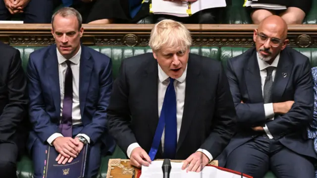 Boris Johnson, a man wearing a dark suit and blue tie, speaking into a microphone at the House of Commons