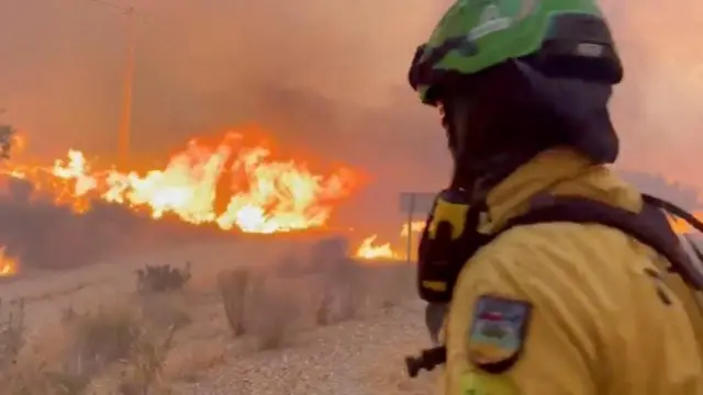 An emergency worker dressed in yellow work wear, wearing a green helmet and breathing equipment looks on as a fire burns nearby on dry land