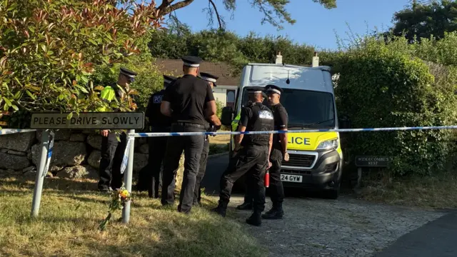 Police men and a van outside a home, a cordon remains in place and a sign says please drive slowly.