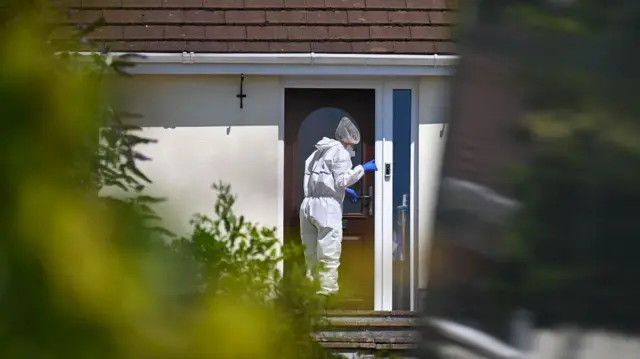 A forensics analyst in a white jumpsuit and blue latex gloves enters Widdecombe's home.