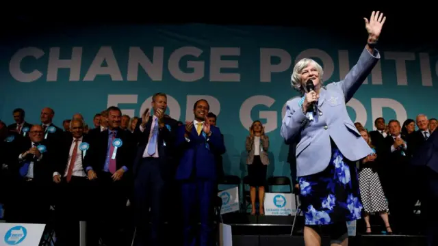 Ann Widdecombe holds her hand up as she delivers a speech at a Brexit Party event. She is wearing a blue blazer. Other Brexit Party members can be seen clapping behind her