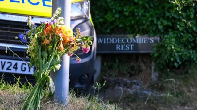 Some flowers are propped up against a pole. A sign saying 'Widdecombe's Rest' could be seen in the background next to a police van