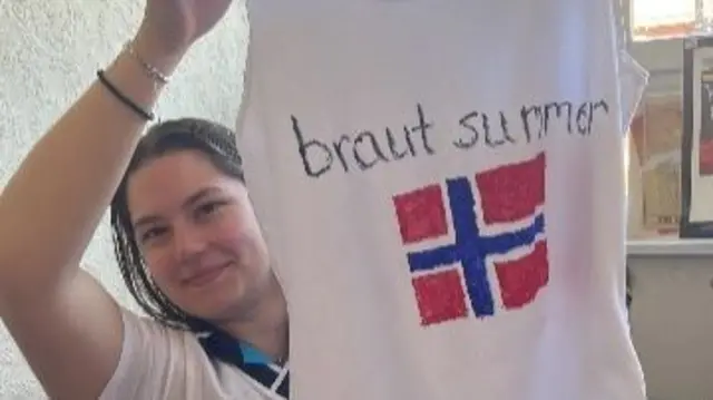 Hanna holds up a handmade vest featuring the Norway flag and 'Braut Summer'