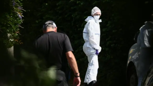 A man in a forensics white uniform is seen at Ann Widdecombe's home. He is stood next to a hedge and a police officer can be seen in front of him