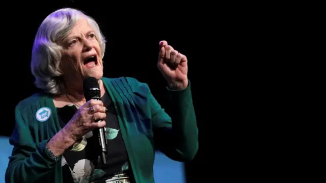 Former Conservative Party MP Ann Widdecombe speaks during a rally. She is holding a microphone and wearing a green cardigan
