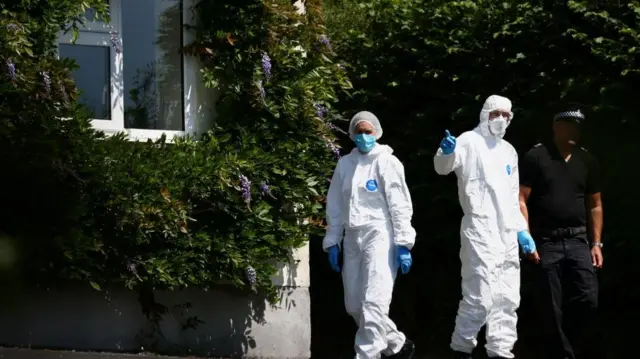 Two forensic workers stand outside the property next to a police officer in uniform