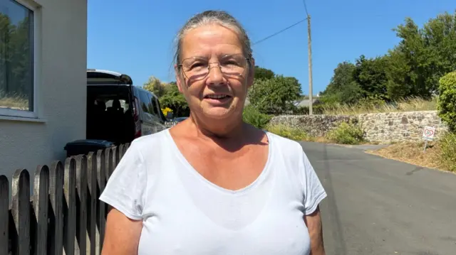 A woman in a T-shirt stands facing the camera, standing on a village street.