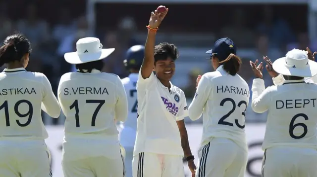 Kranti Gaud (centre), wearing whites, smiles and holds the red cricket ball in her right hand above her head, surrounded by applauding team-mates (two on each side, wearing white and facing away from the camera)