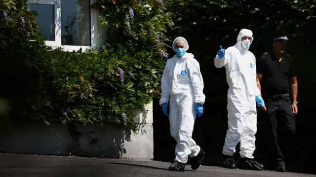 Members of forensics walk outside Ann Widdecombe's home. A police officer wearing a dark uniform is seen next to the forensics officers - who are wearing white uniforms with bags over their shoes. They are also wearing blue surgical masks and gloves