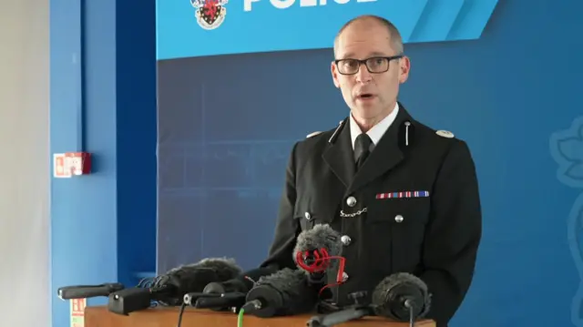 Matt Longman, dressed in a police uniform, speaks behind a bank of microphones.
