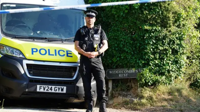A police officer stands in front of a marked police van next to a sign reading 'Widdecombe's Rest'