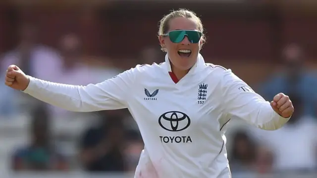 Sophie Ecclestone, wearing a white England shirt and sunglasses, smiles and holds her arms outstretched in celebration