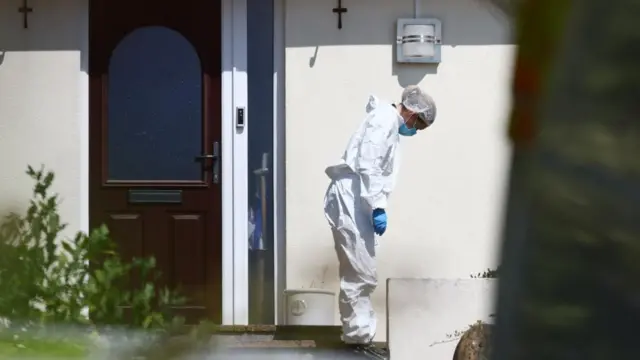 A forensics officer looks down close to a step near a door to Ann Widdecombe's home. She is wearing a hair net, surgical mask, gloves and a white uniform
