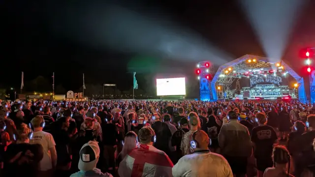 Festival goers at 2000trees festival watch England on big screen using silent disco headphones