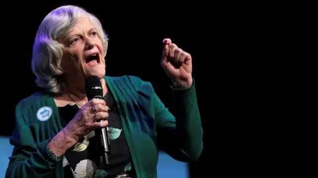 Ann Widdecomb is picturd speaking at an event, holding a microphone and wearing a green jacket over a floral patterned top.