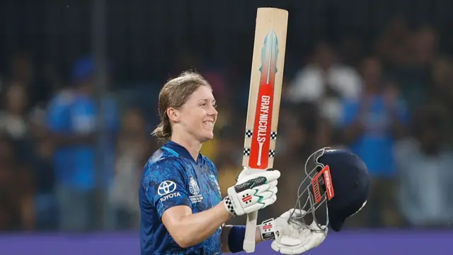 Heather Knight, wearing a blue England shirt and white batting gloves, smiles and raises her bat in her right hand while holding a blue helmet in her left