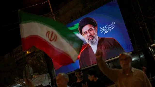 Mojtaba Khamenei pictured on screen during a rally in Tehran, supporters hold up an Iranian flag