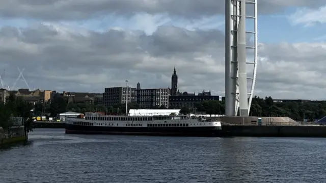 A ship on water, moored near a Science Centre in Glasgow, with large poles and buildings in the background