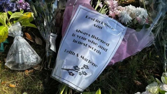 A note lies on top of a bunch of flowers, it reads: "Rest in peace, Ann. Always listened to your wise and honest words. Keith and Marlene".