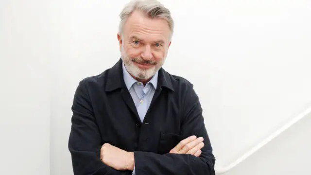 Sam Neill smiling and wearing a dark blue jacket against a white background