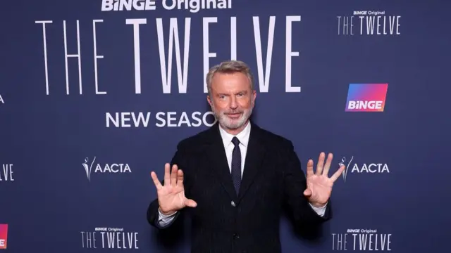 Sam Neill standing with his hands raised, dressed in a dark suit