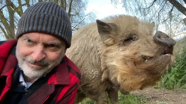 Sam Neill wearing a wolly hat and posing for a selfie with one of his pigs