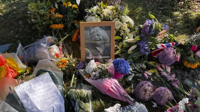 Dozens of bunches of flowers sit on the side of a road, with a photograph of Ann Widdecombe in a brown wooden flame.