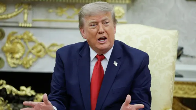 Donald Trump, wearing a dark blazer and red tie, gestures with his hands as he sits on a cream coloured chair in the Oval Office.