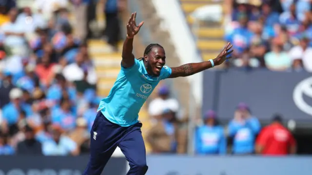 Jofra Archer, wearing a bright blue England shirt and dark blue trousers, runs with both arms in the air and a grin
