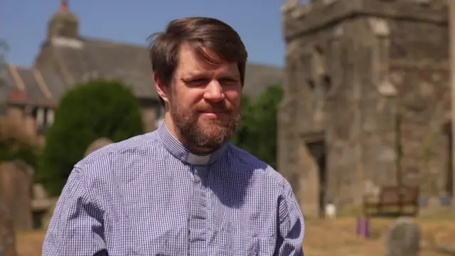 Phill Parker has brown hair and a beard and wears a blue and white checkered shirt and clerical collar.