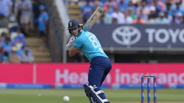 Jacob Bethell, wearing a bright blue shirt, dark blue trousers and helmet, holds his bat over his right shoulder and looks over his front left shoulder to follow the trajectory of the ball