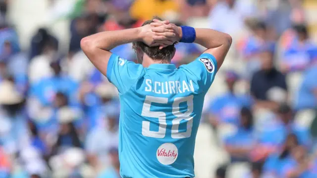 Sam Curran, photographed from behind, wearing a light blue England shirt with S Curran 58 on the back, stands with his hands behind his hed