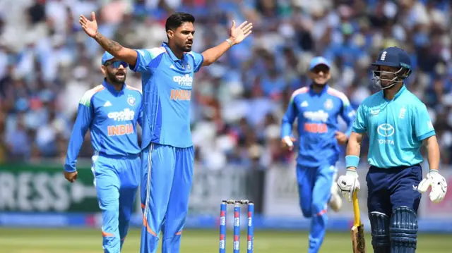 Bowler Gurnoor Brar, wearing all blue, stares at Ben Duckett (right), wearing a bright blue shirt and dark blue trousers/helmet, with his arms outstretched