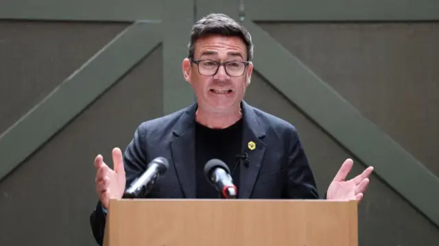 Andy Burnham stands behind a lectern, wearing a dark coloured blazer and a black t-shirt underneath.
